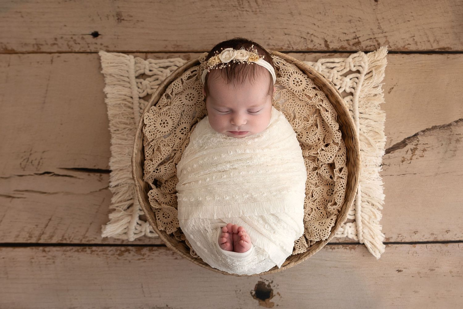 Sleeping newborn wrapped in white swaddle lying in round basket with textured pillows on wooden floor.