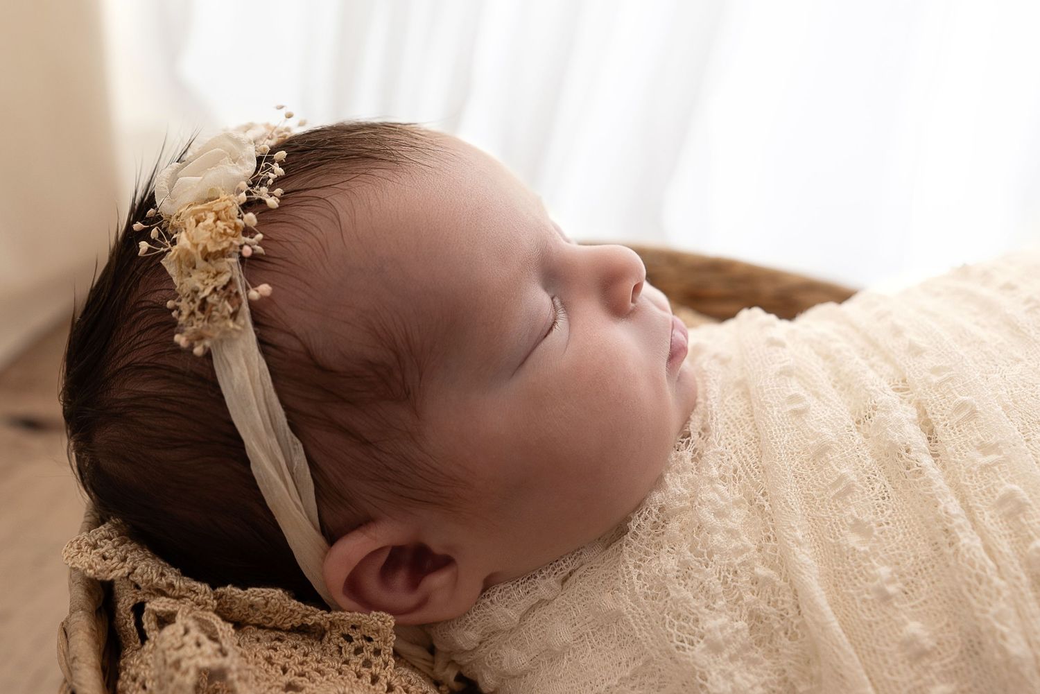 A sleeping baby wearing a delicate lace dress and floral headband rests peacefully against cream-colored fabric.