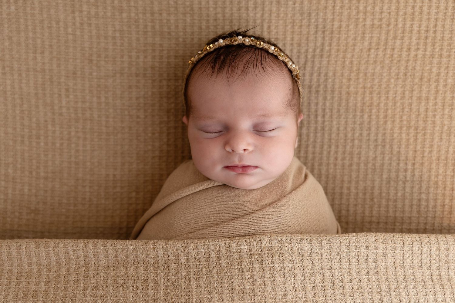 Sleeping newborn baby wrapped in beige fabric wearing delicate headband against neutral textured background.
