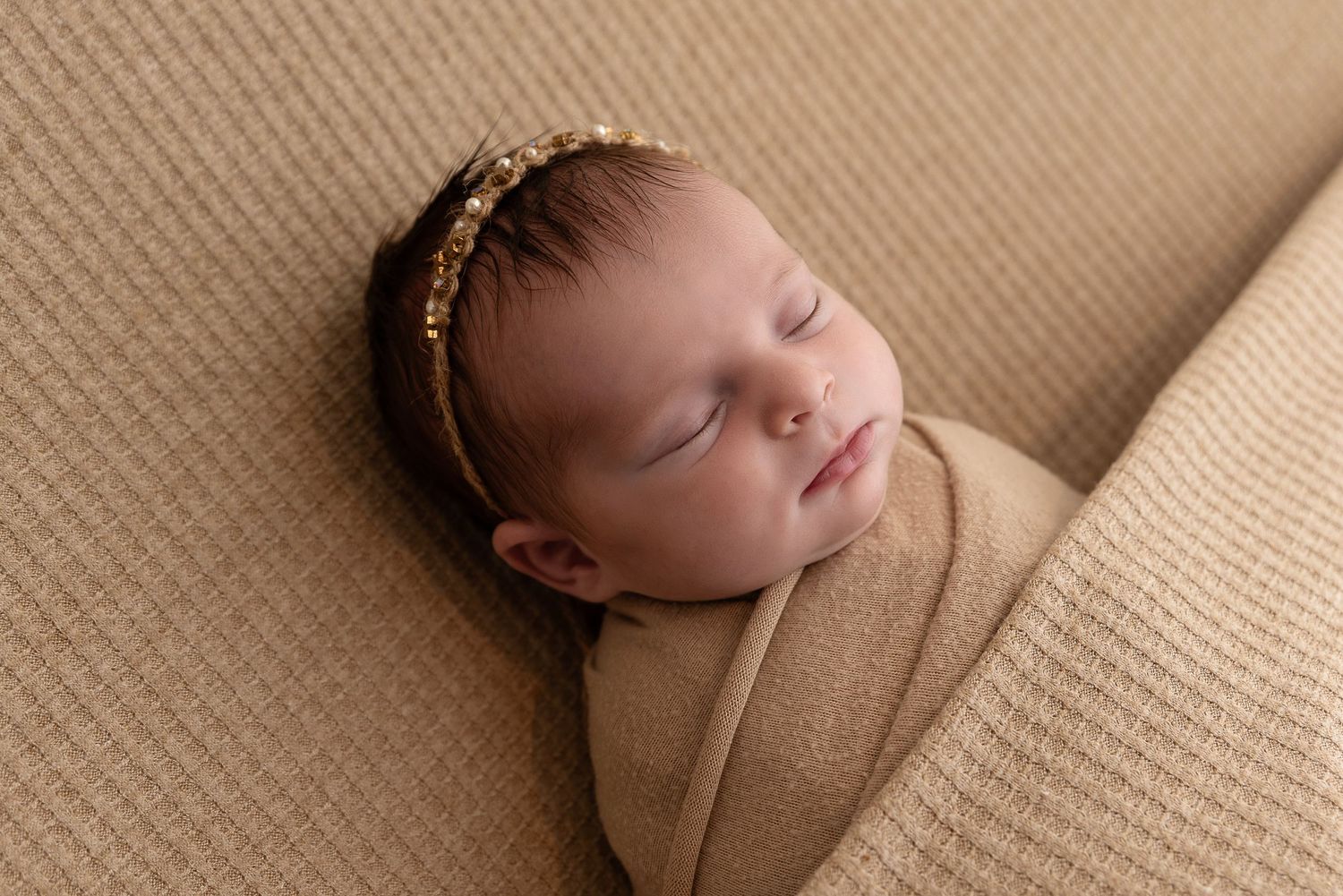 Peaceful sleeping infant in beige wrap against neutral textured backdrop wearing delicate headpiece.