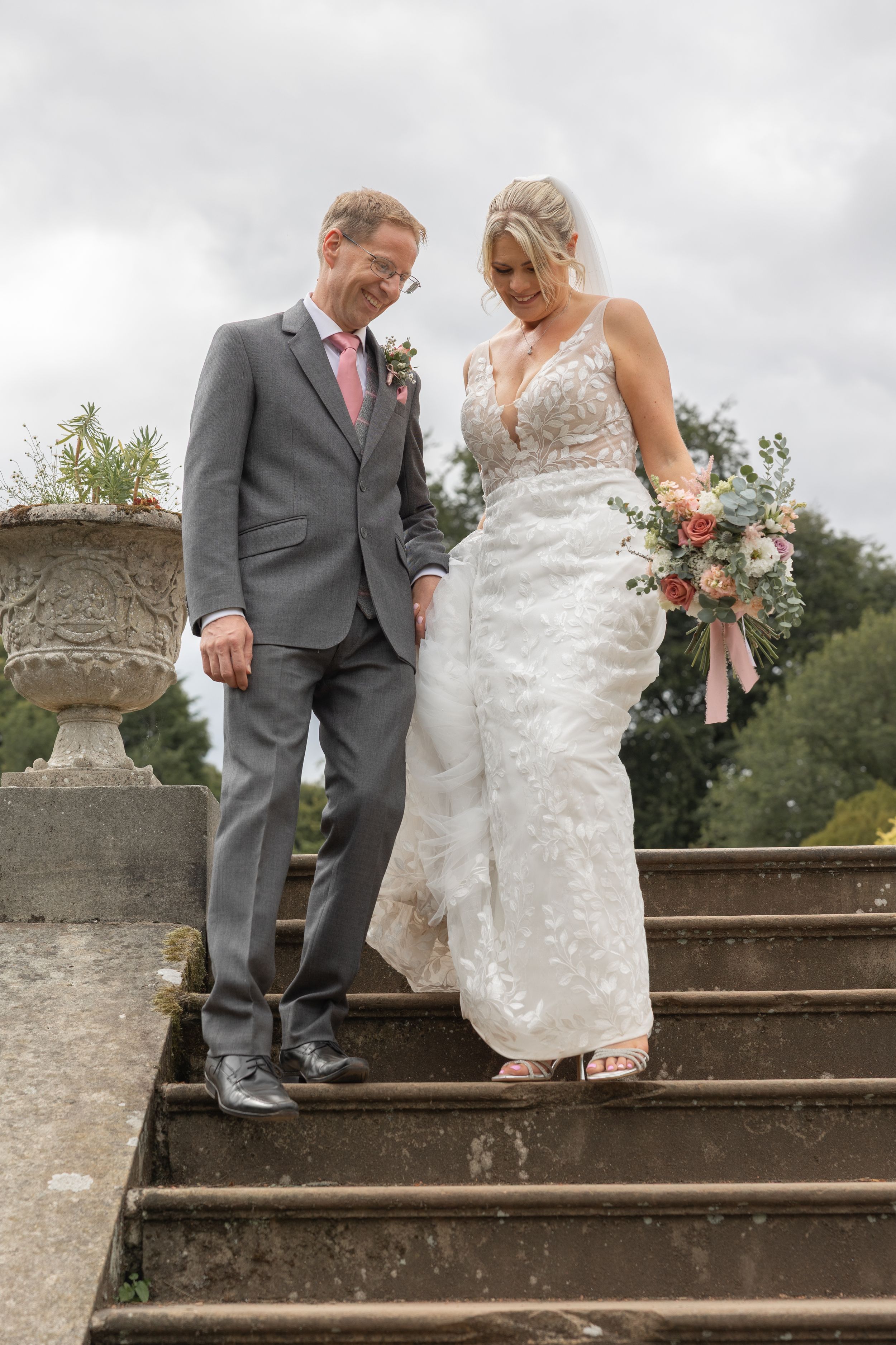 A couple in wedding attire ascends stone steps together in a romantic outdoor garden setting.
