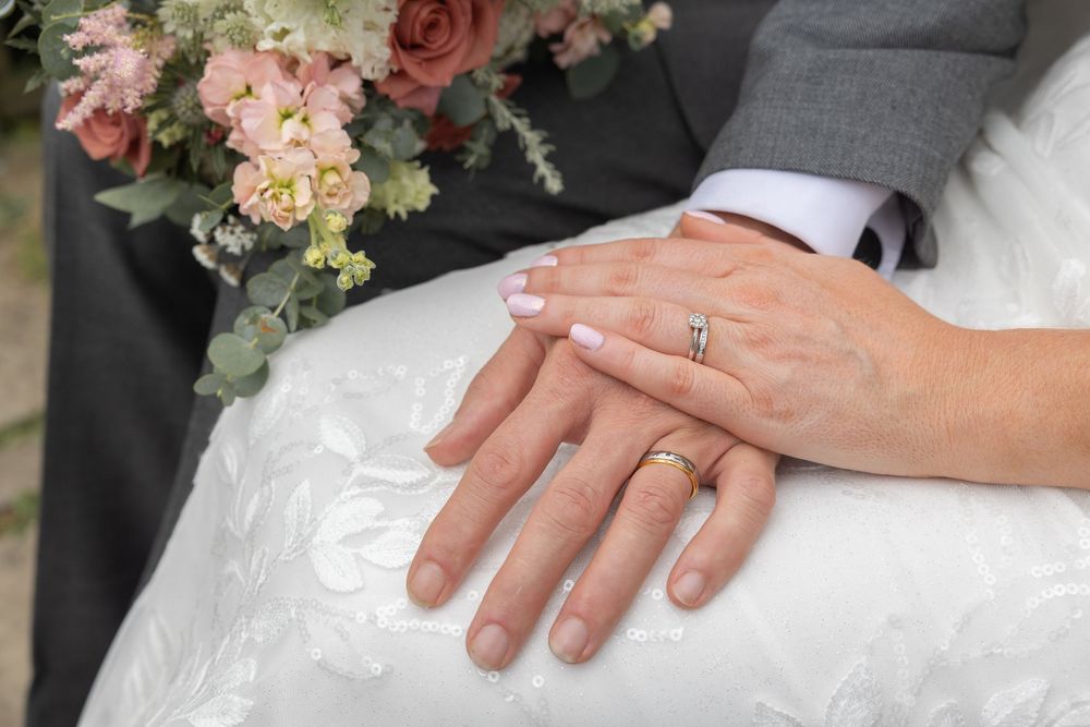 Wedding hands with rings resting on white dress fabric next to pink and peach floral bouquet.
