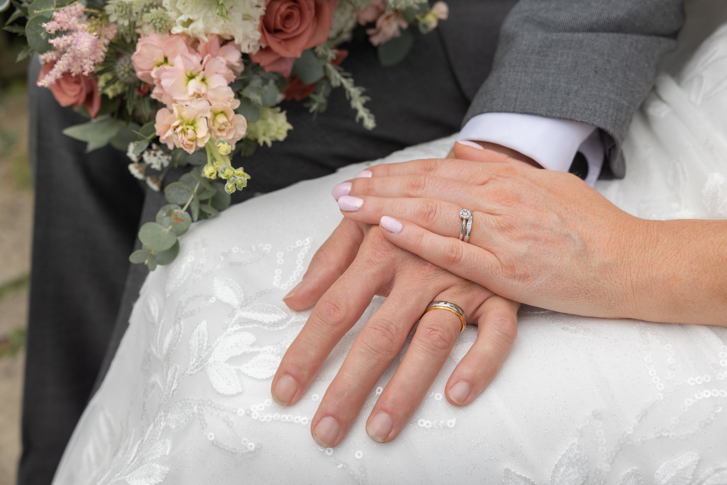 Wedding hands with rings resting on white dress fabric next to pink and peach floral bouquet.
