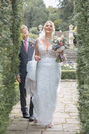 A bride in a white lace gown walks along a stone path surrounded by greenery during an outdoor wedding ceremony.
