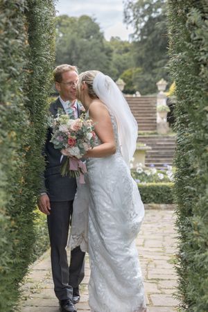 A romantic wedding couple shares a tender moment in a garden pathway lined with greenery and stone steps.