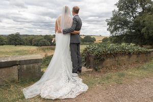 A couple in wedding attire embraces while overlooking a scenic countryside landscape on an overcast day.