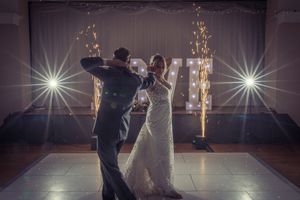 Wedding couple dances in a dark ballroom illuminated by dramatic spotlights and sparklers creating a magical atmosphere.