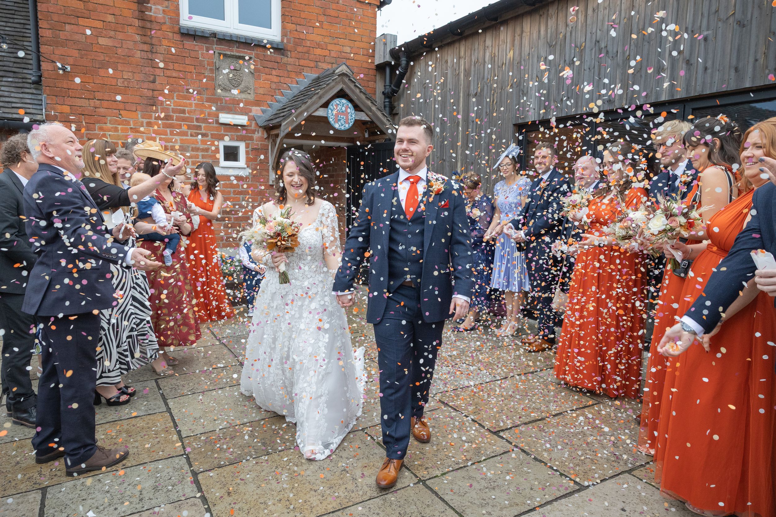 Wedding guests throw confetti in celebration outside a rustic brick building with a festive autumn display.