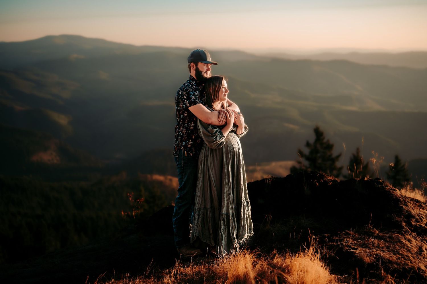 A couple embraces on a mountaintop during golden hour with rolling hills in the background.