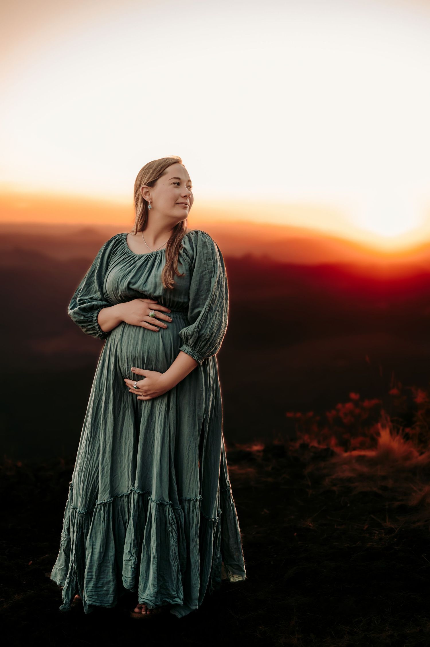 Expectant mother in green dress poses against sunset mountain landscape.