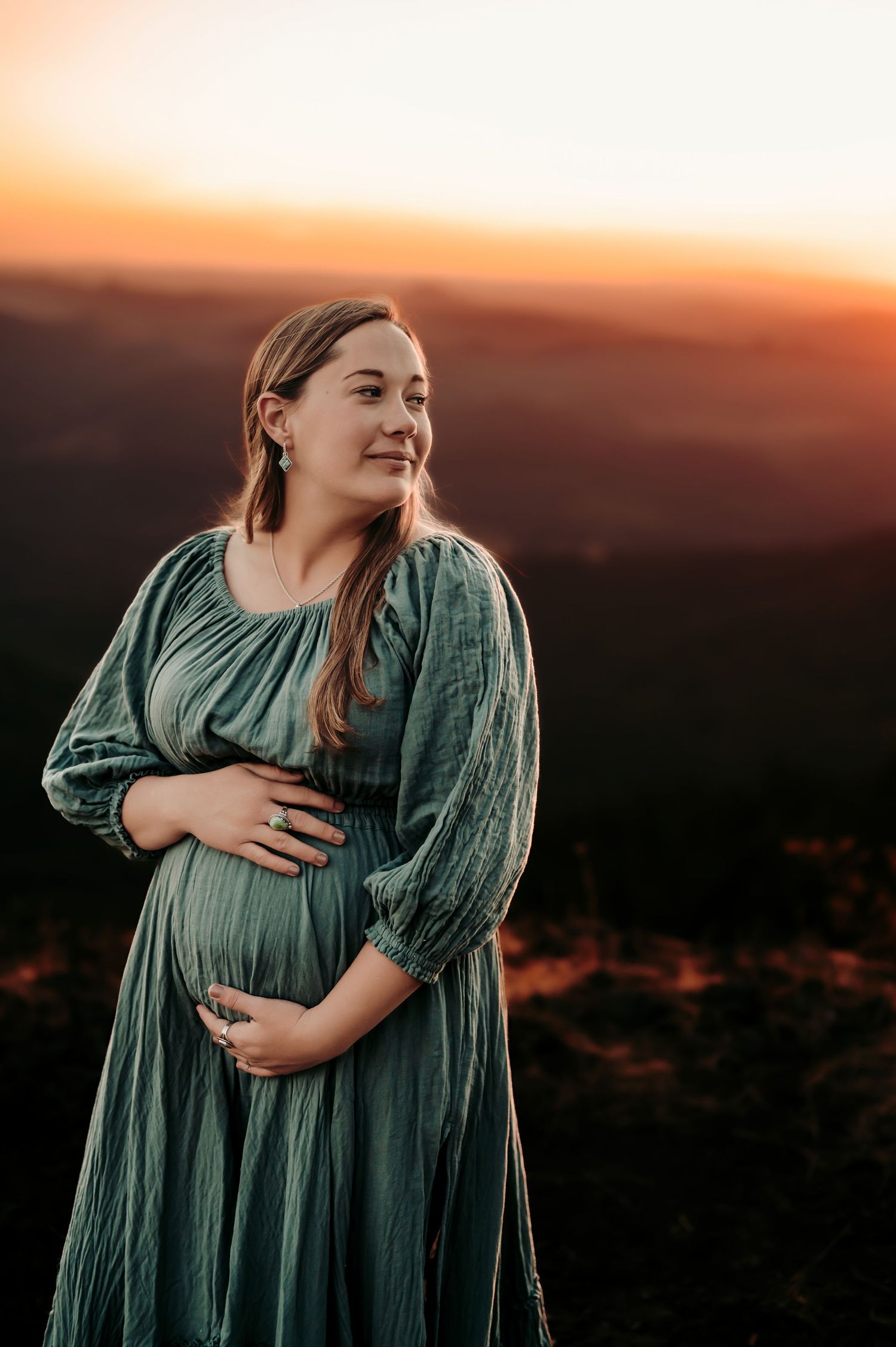 Pregnant woman in sage green dress smiles during sunset mountain photoshoot.