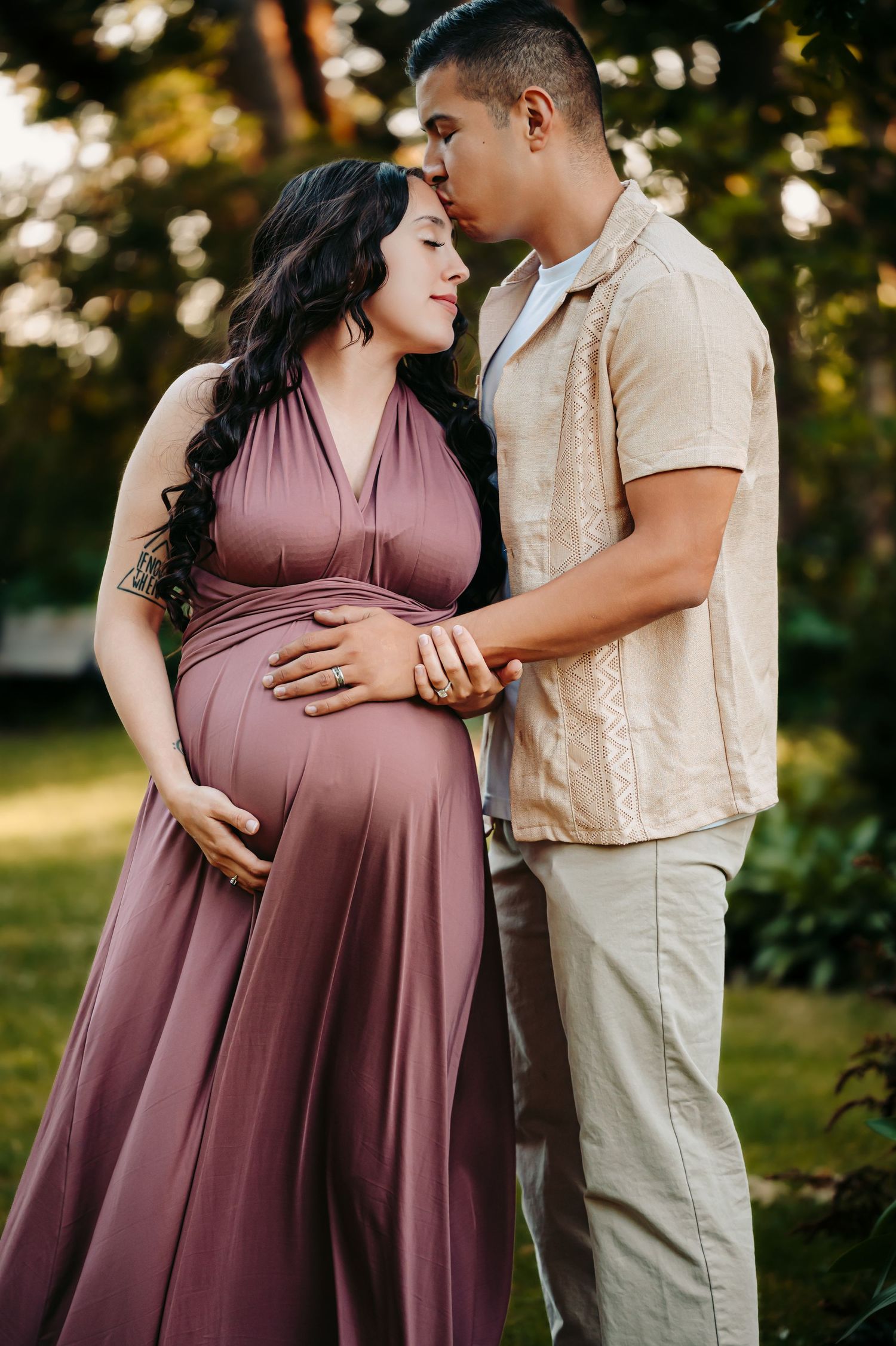 Romantic maternity portraits in a mauve dress against lush greenery at sunset.
