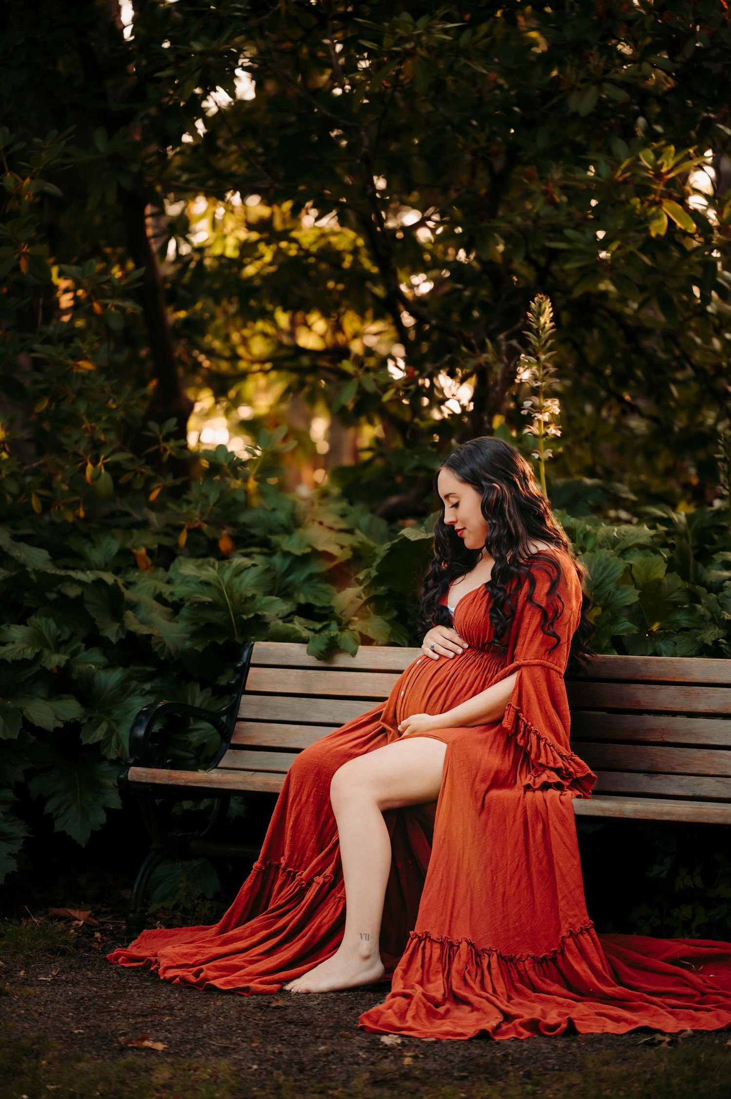 Dramatic red dress maternity photos on a wooden bench surrounded by lush foliage.