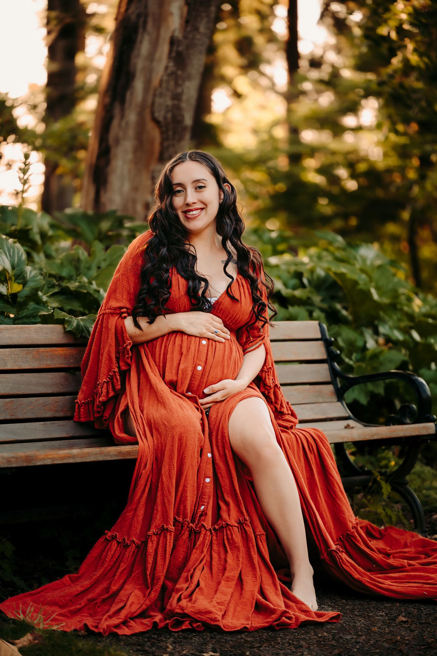 Vibrant rust-colored dress maternity portraits on a garden bench with natural backdrop.