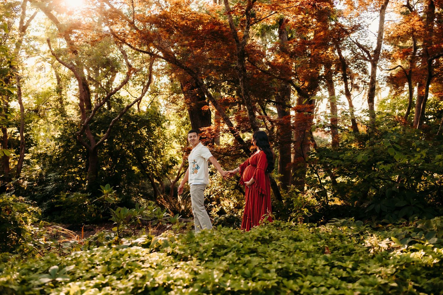 Romantic maternity photoshoot in a sunlit forest with golden light filtering through trees.