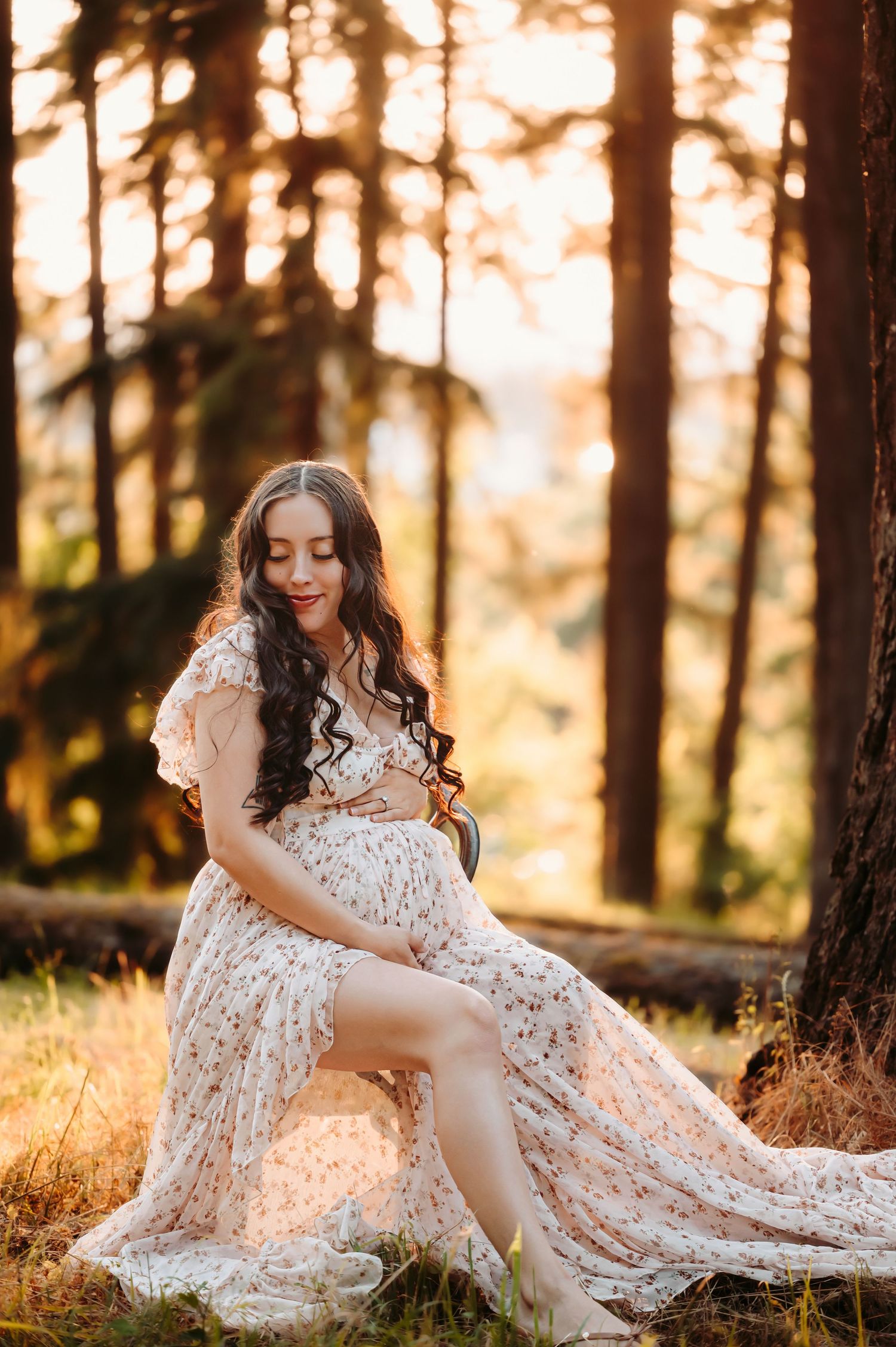 Ethereal maternity portrait of woman in lace dress sitting among pine trees at sunset.