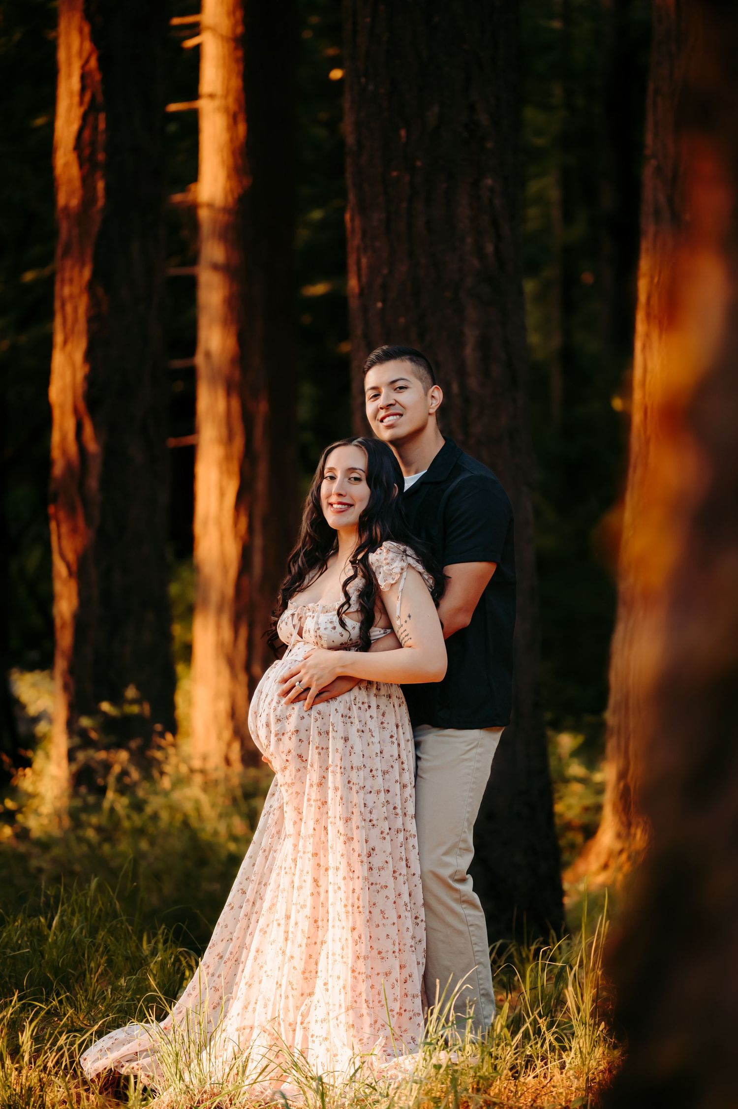 Golden hour maternity photos with couple embracing among tall pine trees.