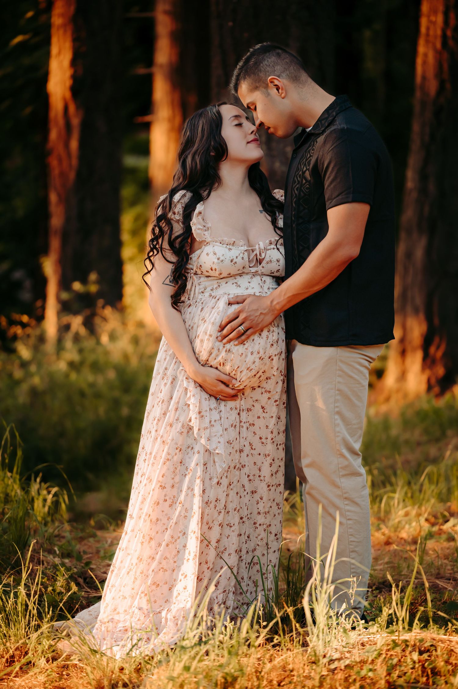 Romantic maternity photoshoot in a sunlit forest with couple embracing in flowing floral dress at sunset.