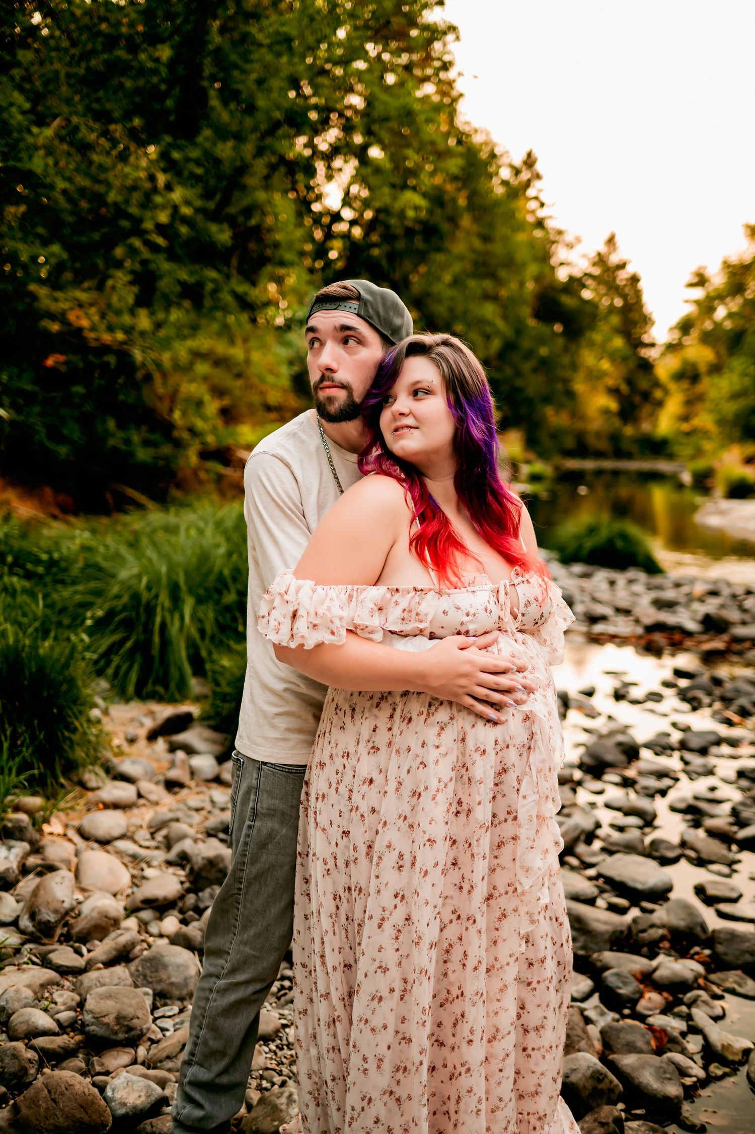 Couple shares intimate moment on rocky riverbank during sunset maternity session.
