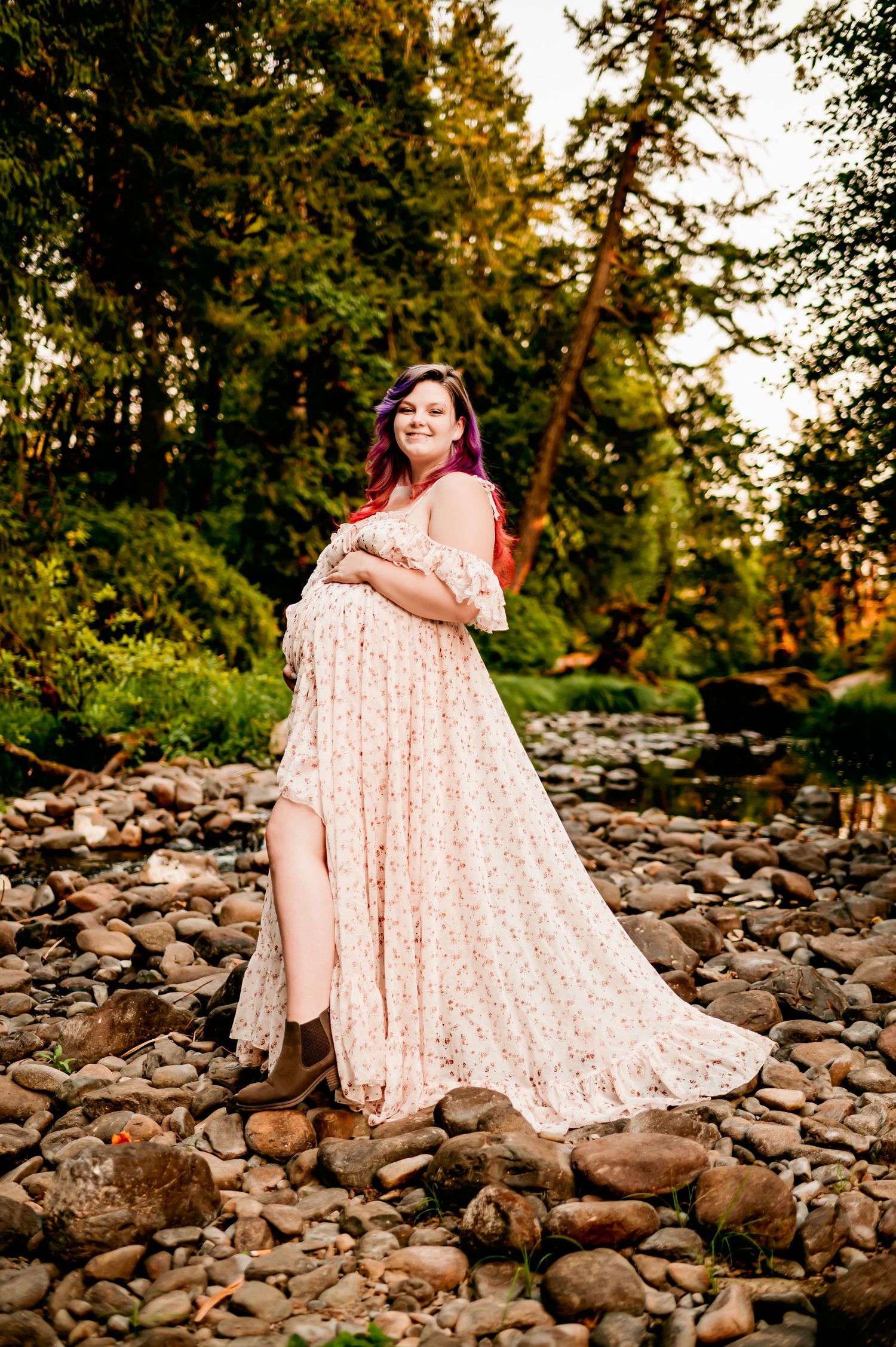 Expectant mother in lace dress poses gracefully on rocky riverbank during golden hour.
