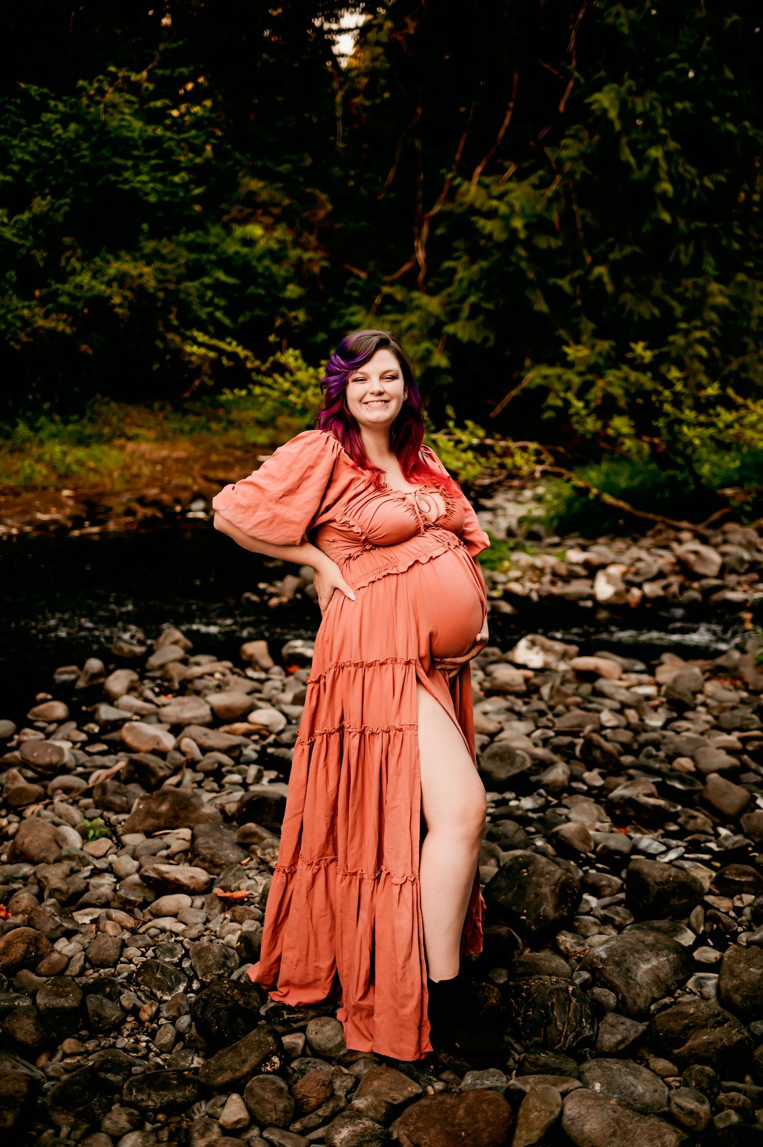 Artistic maternity portraits on stone riverbed with coral dress and natural backdrop.