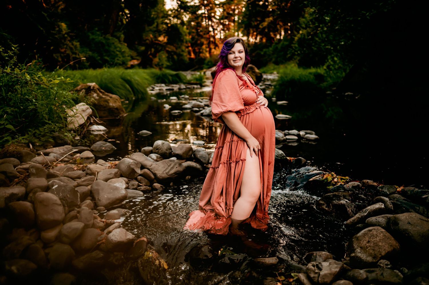 Maternity photography by flowing stream with rocks and forest backdrop at dusk.