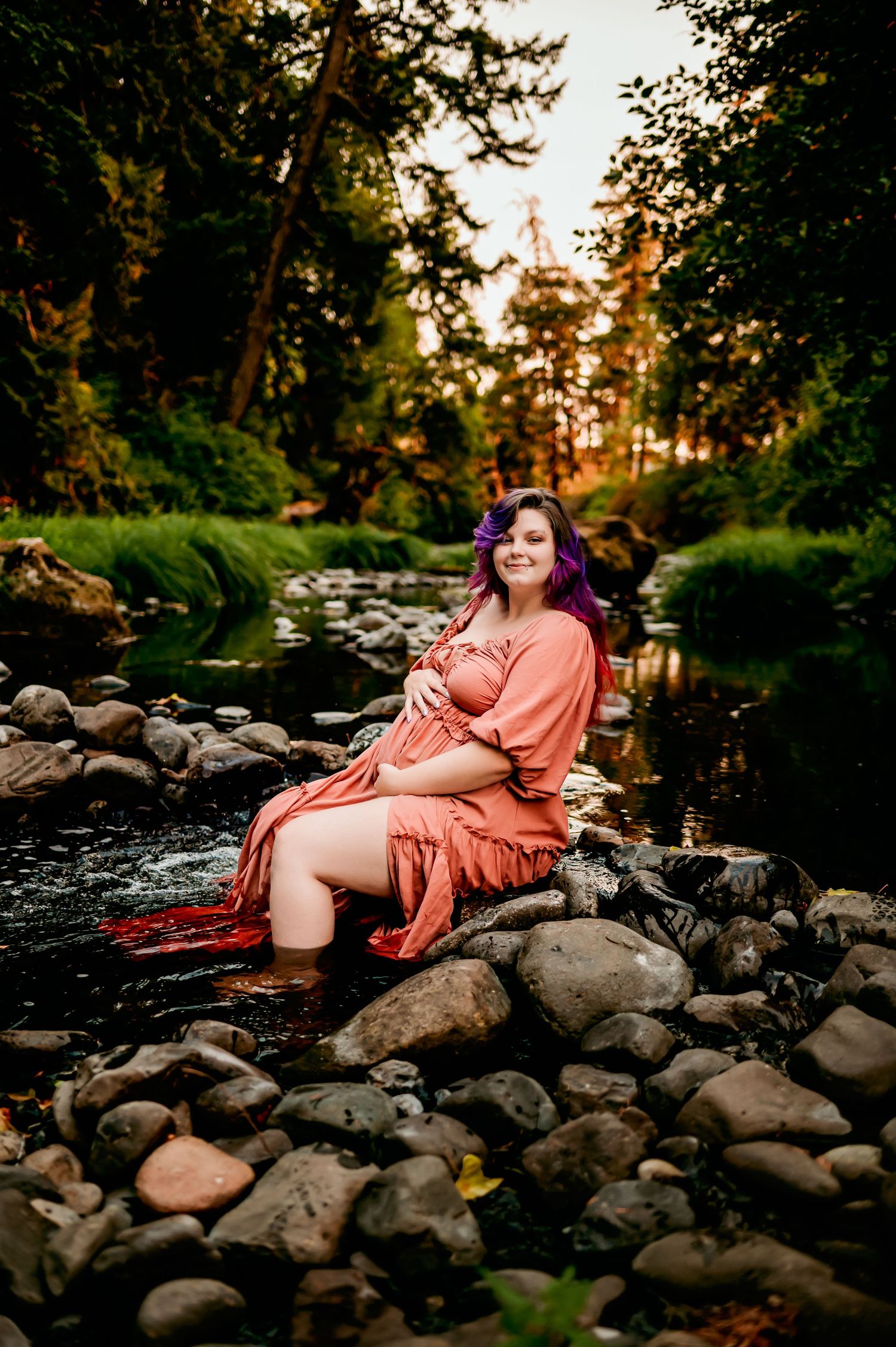 Sunset maternity session by rocky stream with coral dress and natural scenery.