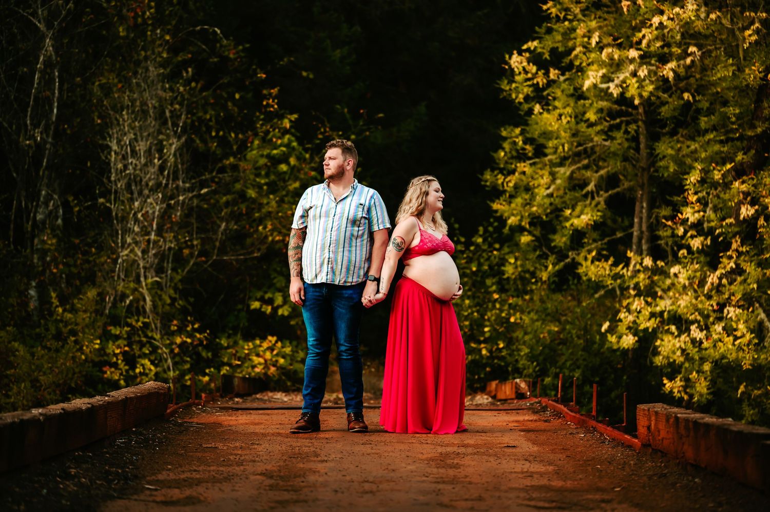 Expecting couple poses on wooden bridge surrounded by autumn foliage.
