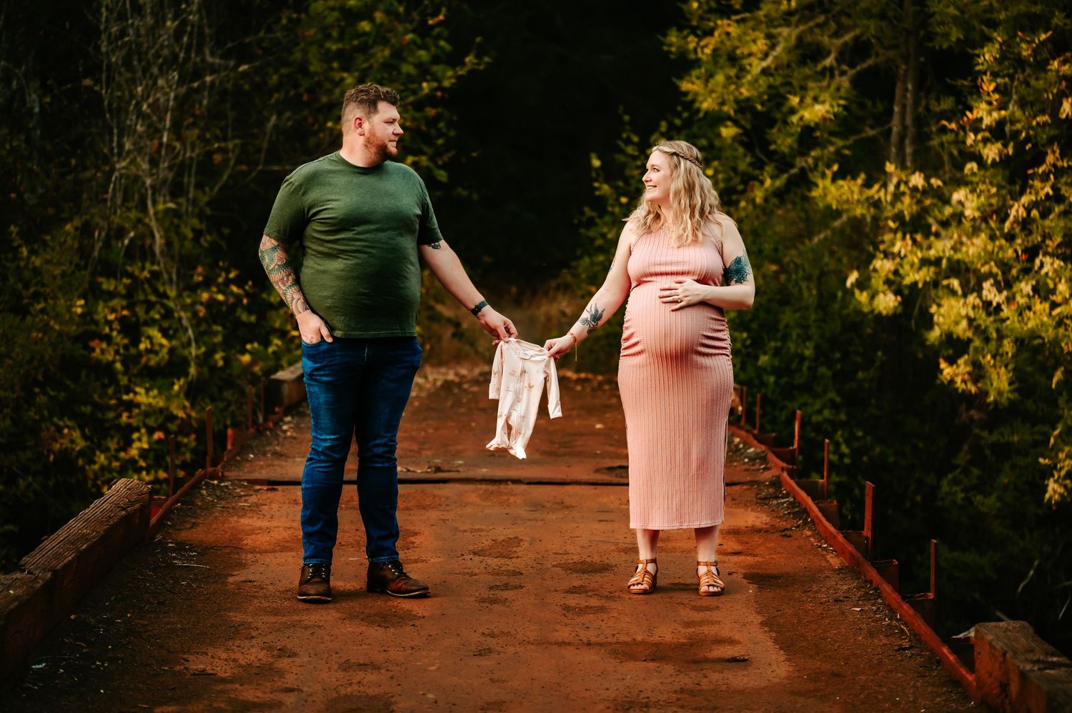 Couple displays baby clothes while standing on wooden bridge in forest setting.