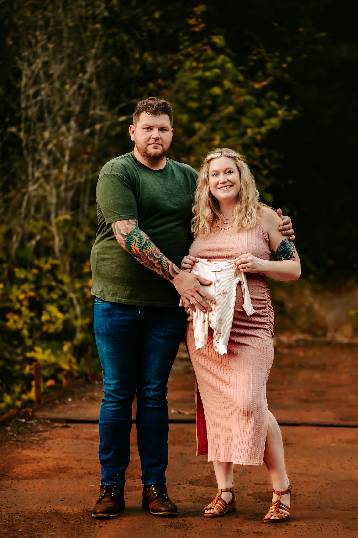 Couple holding baby clothes poses on rustic bridge during maternity photoshoot.