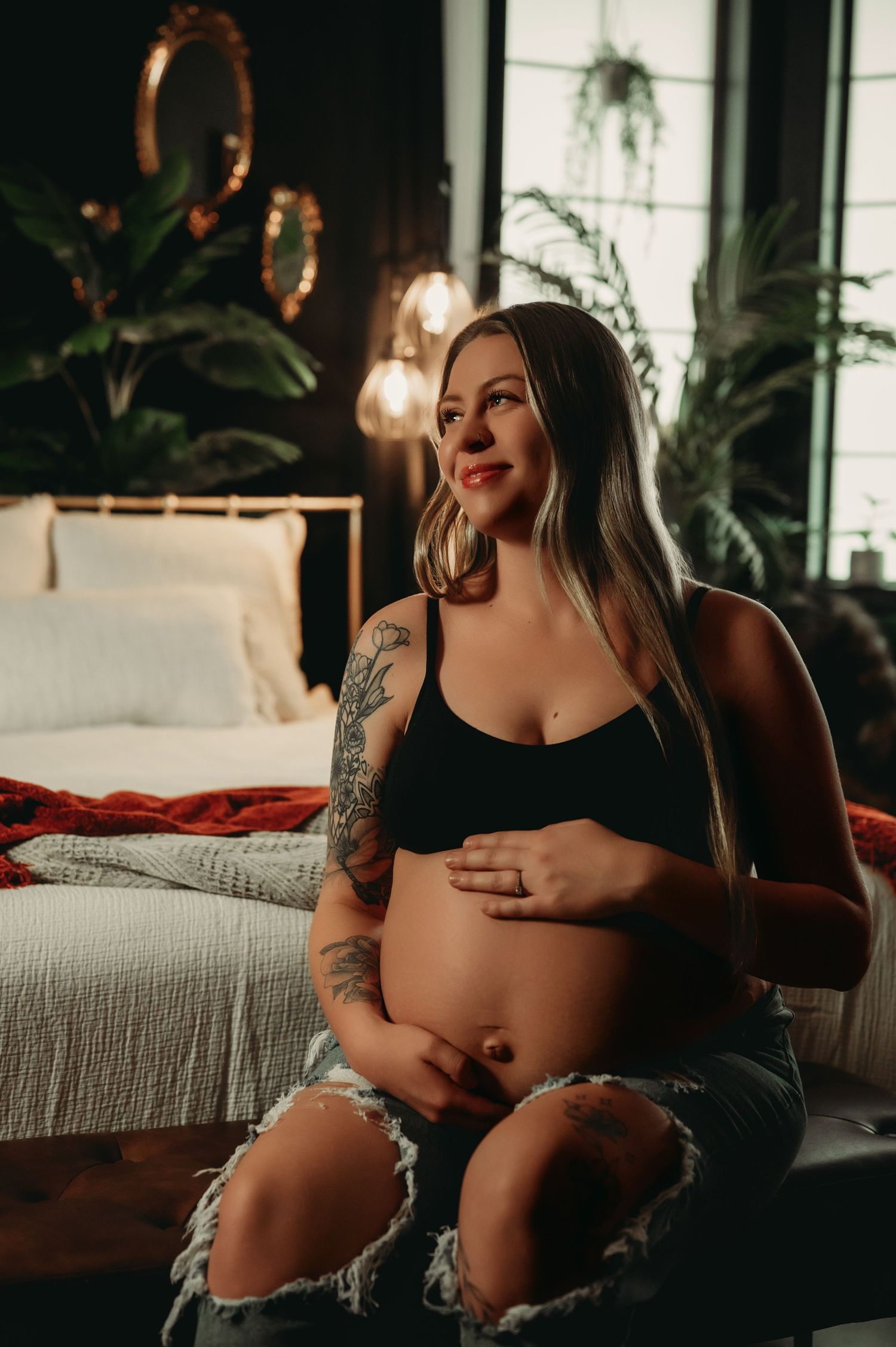 Artistic maternity photoshoot featuring soft lighting and indoor plants in a cozy bedroom setting.