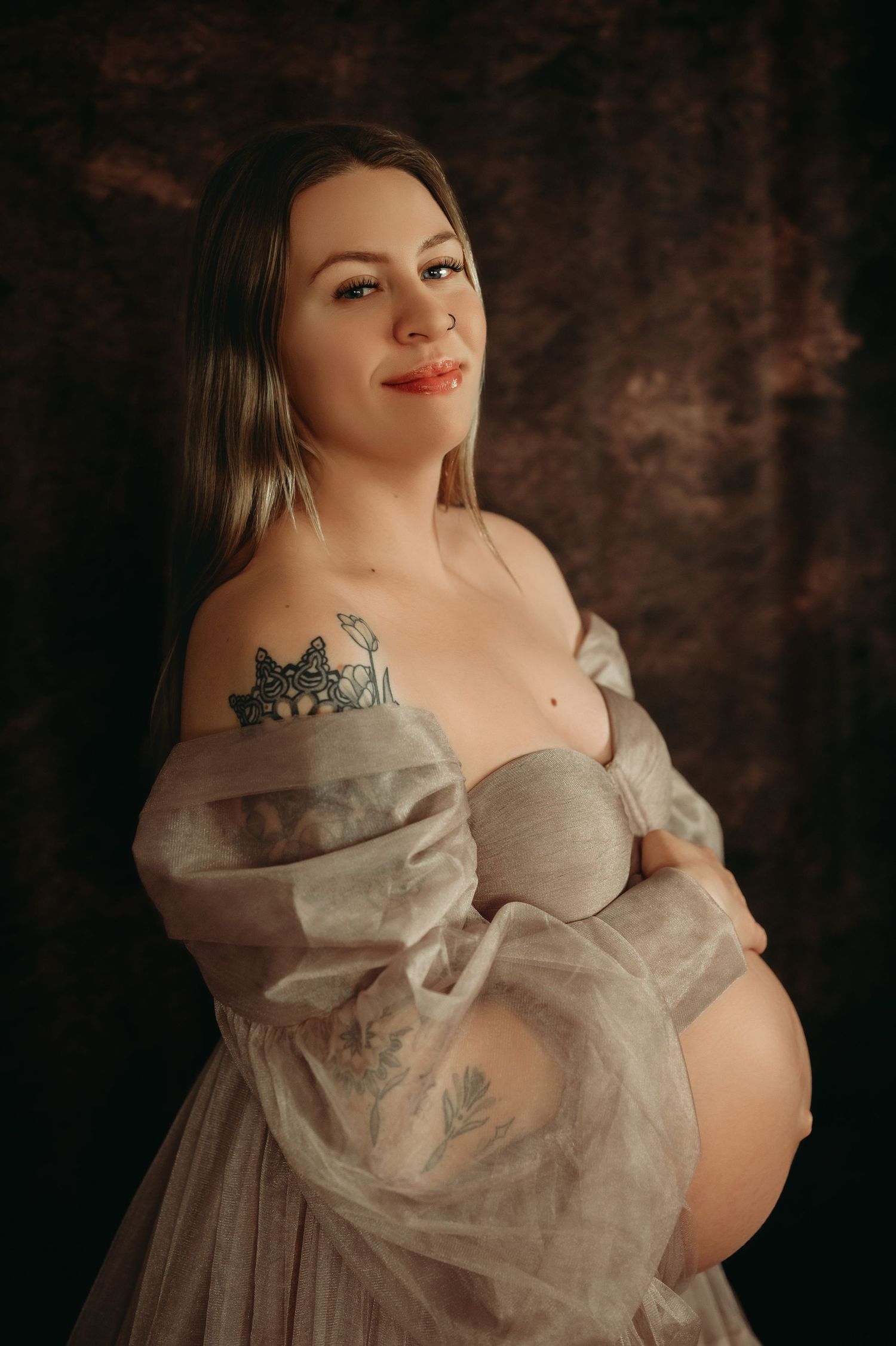 Woman in ethereal off-shoulder tulle gown poses against dark backdrop in romantic maternity photoshoot.