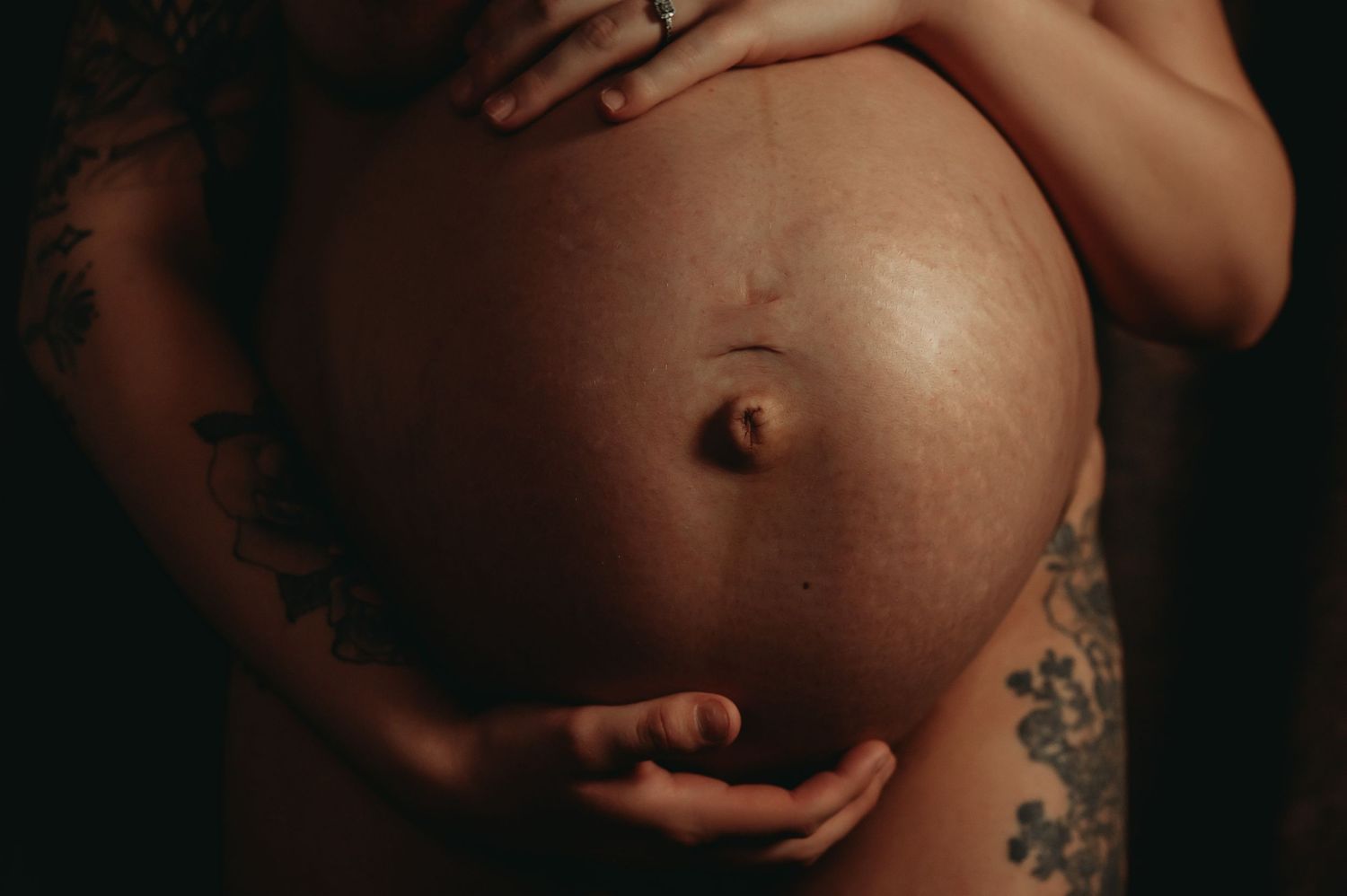 Close-up photo of a pregnant belly with visible tattoo art and hands cradling the bump against dark background.