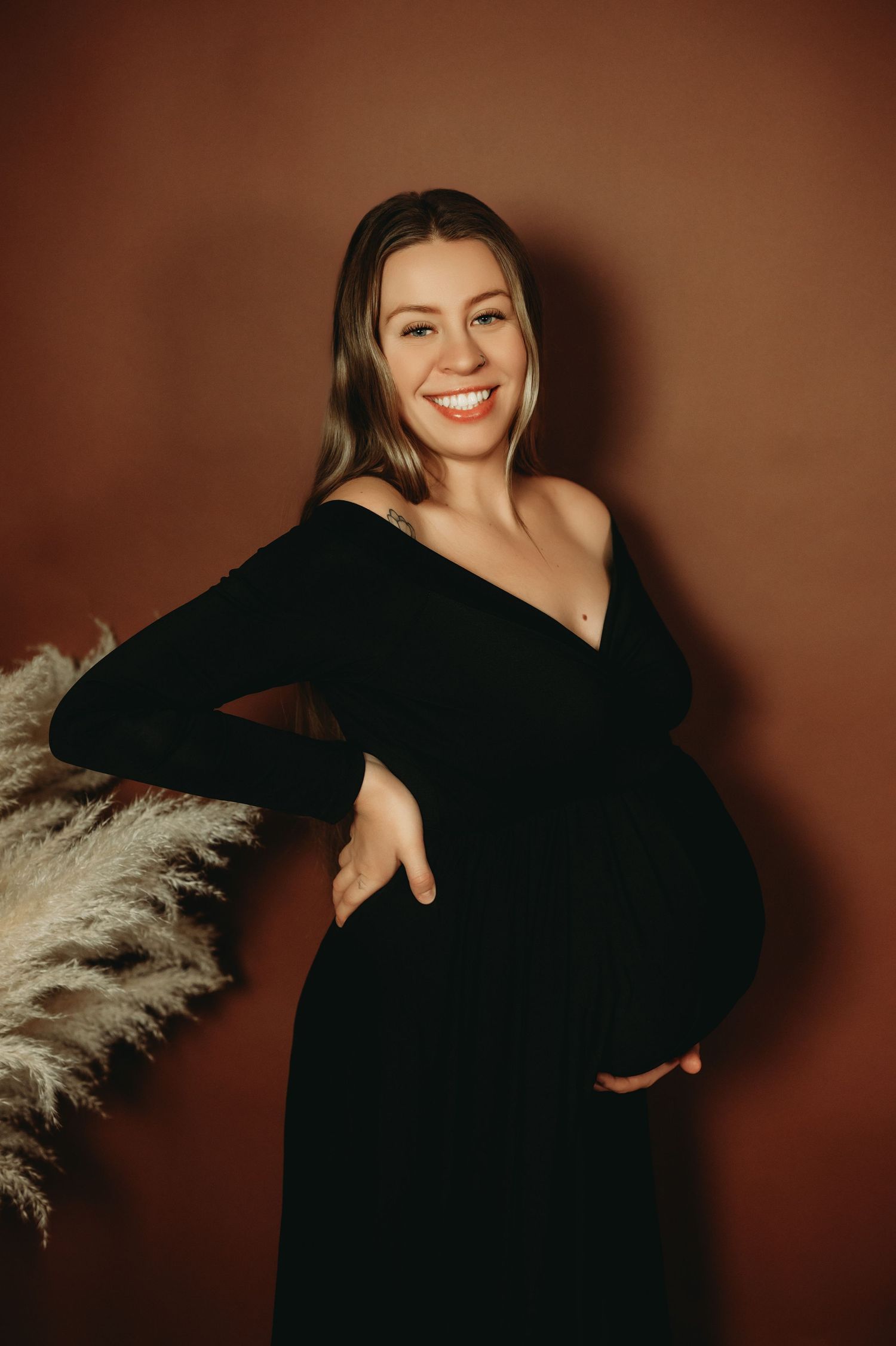 A figure in a black long-sleeve dress poses against a warm brown backdrop with pampas grass decor.