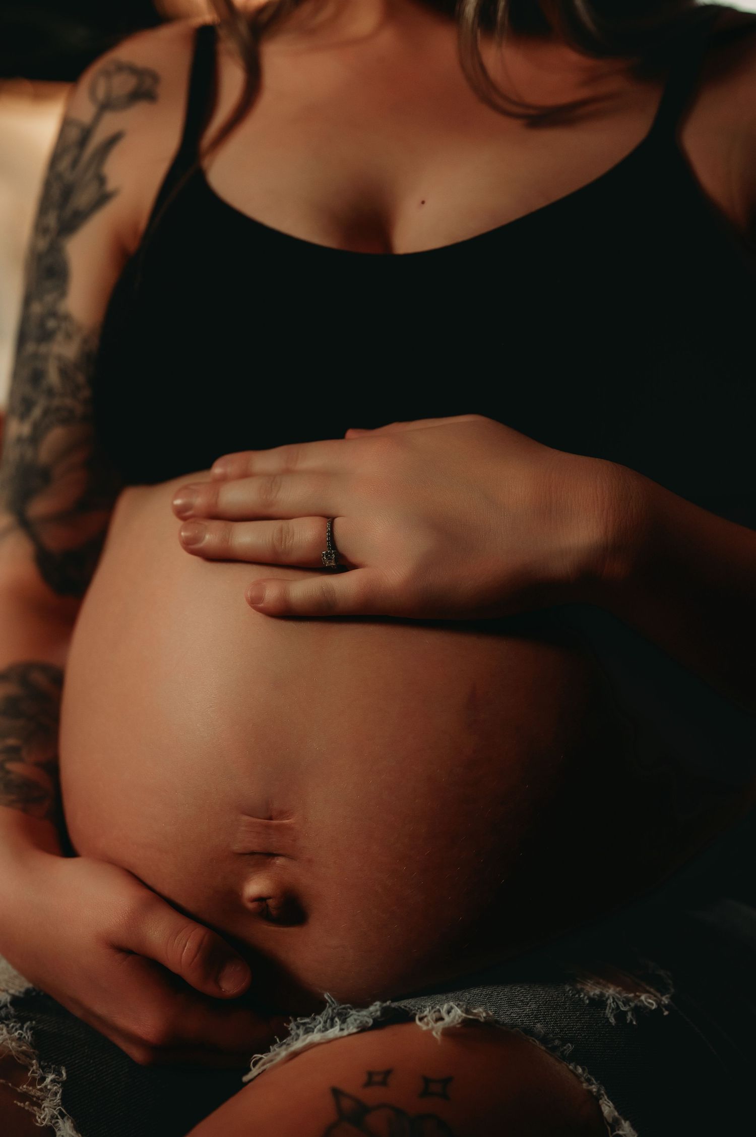 Close-up view of a pregnant belly in black lingerie with tattoos visible against a dark background.