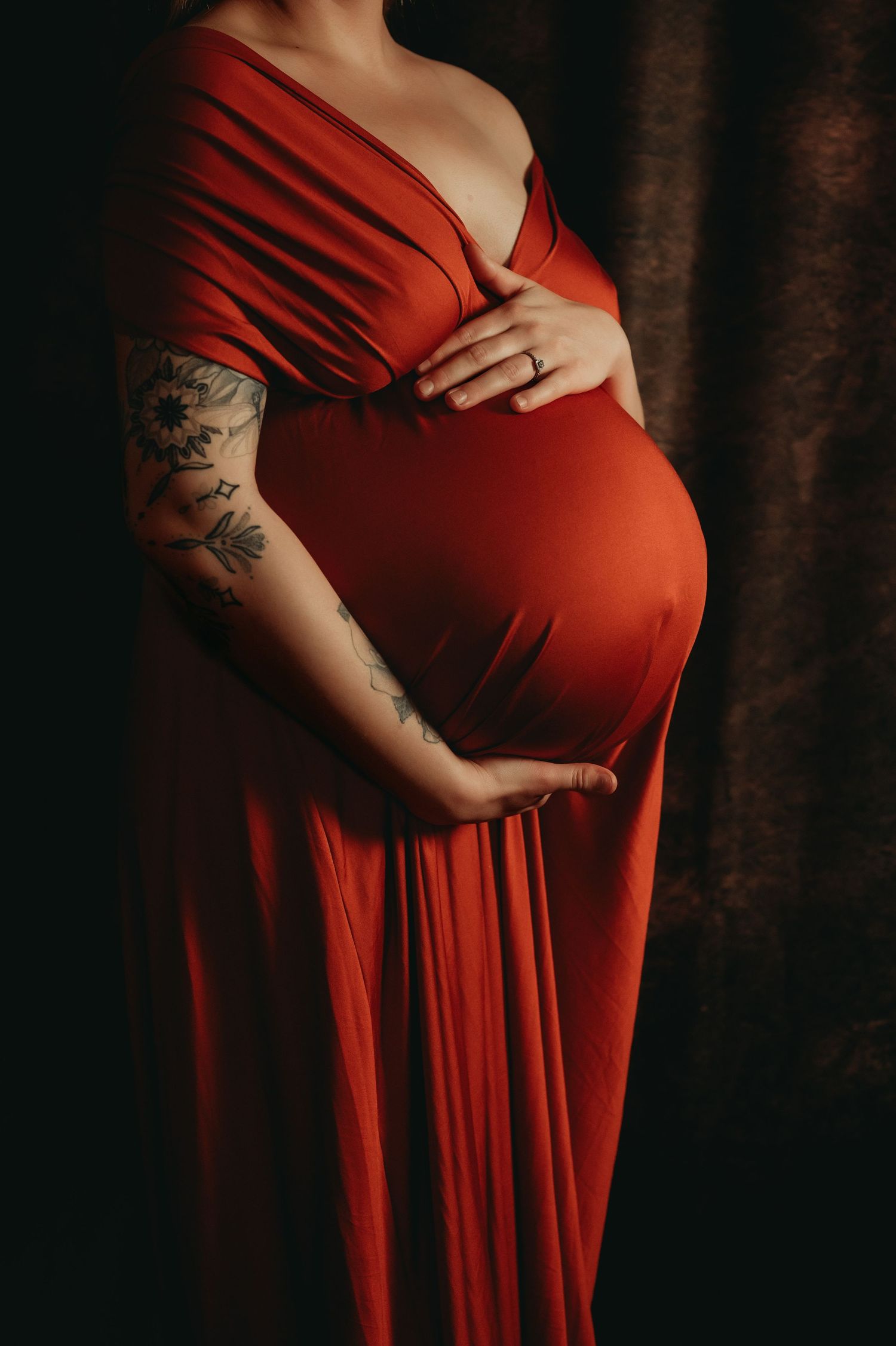 Dramatic maternity portrait in flowing red dress against dark backdrop.
