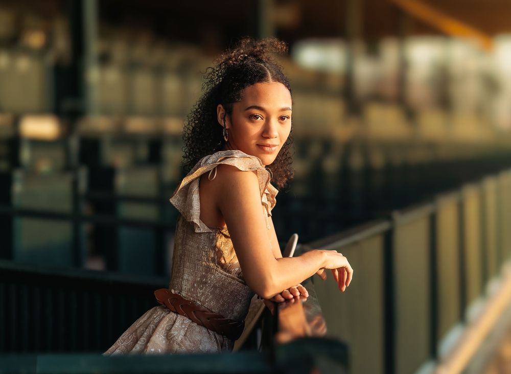 Young woman in a sparkly formal dress leans against a railing during golden hour with warm evening lighting.