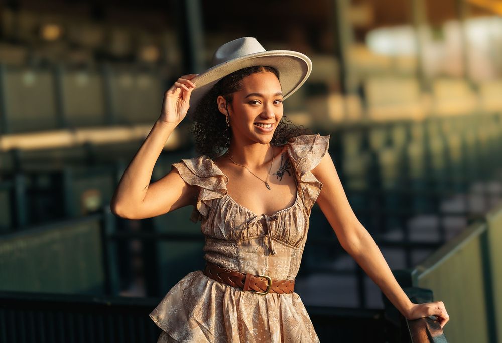 A woman in a vintage-style dress and wide-brimmed hat poses gracefully during golden hour at a rustic outdoor location.