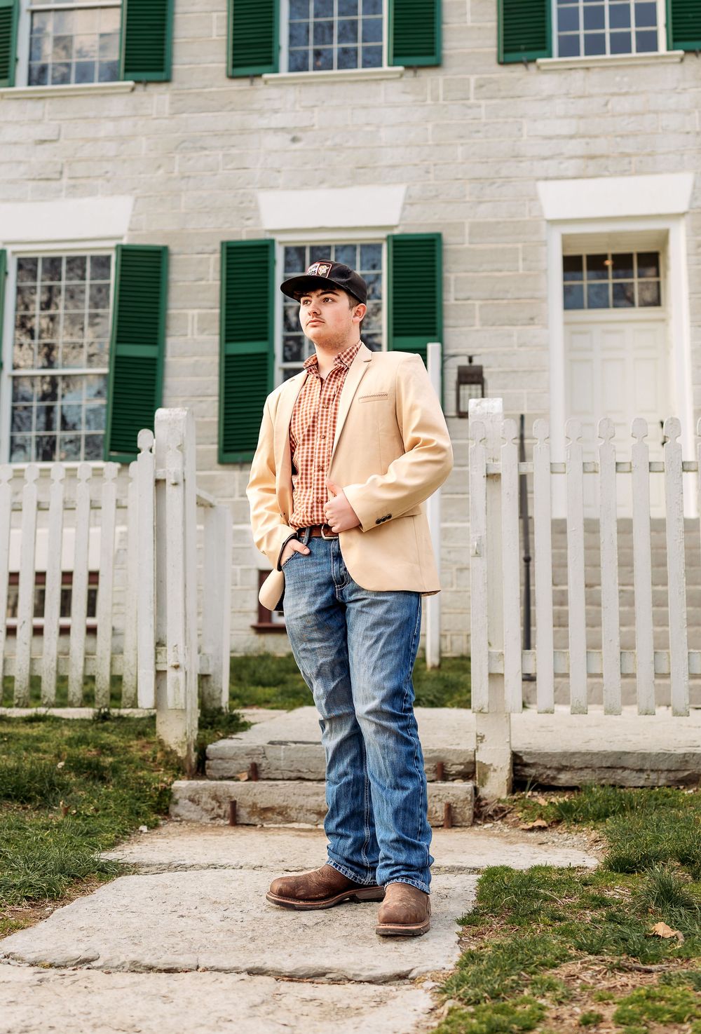 Individual wearing a beige jacket, jeans and boots stands in front of a white house with green shutters and picket fence.