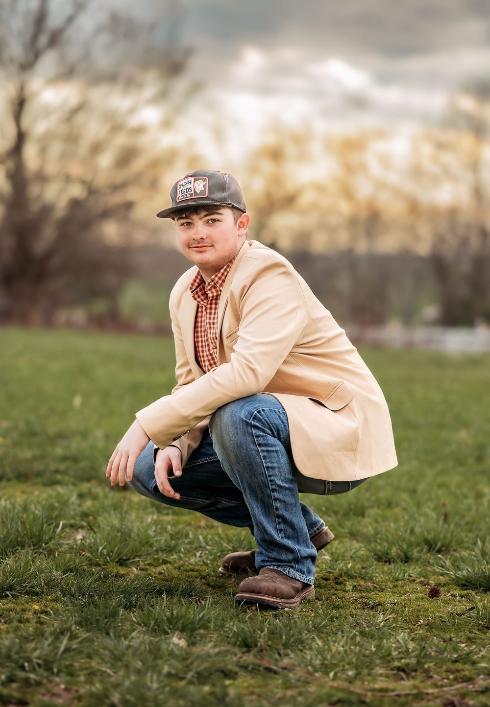 Person in a baseball cap, plaid shirt and beige jacket crouches on grassy field during autumn.