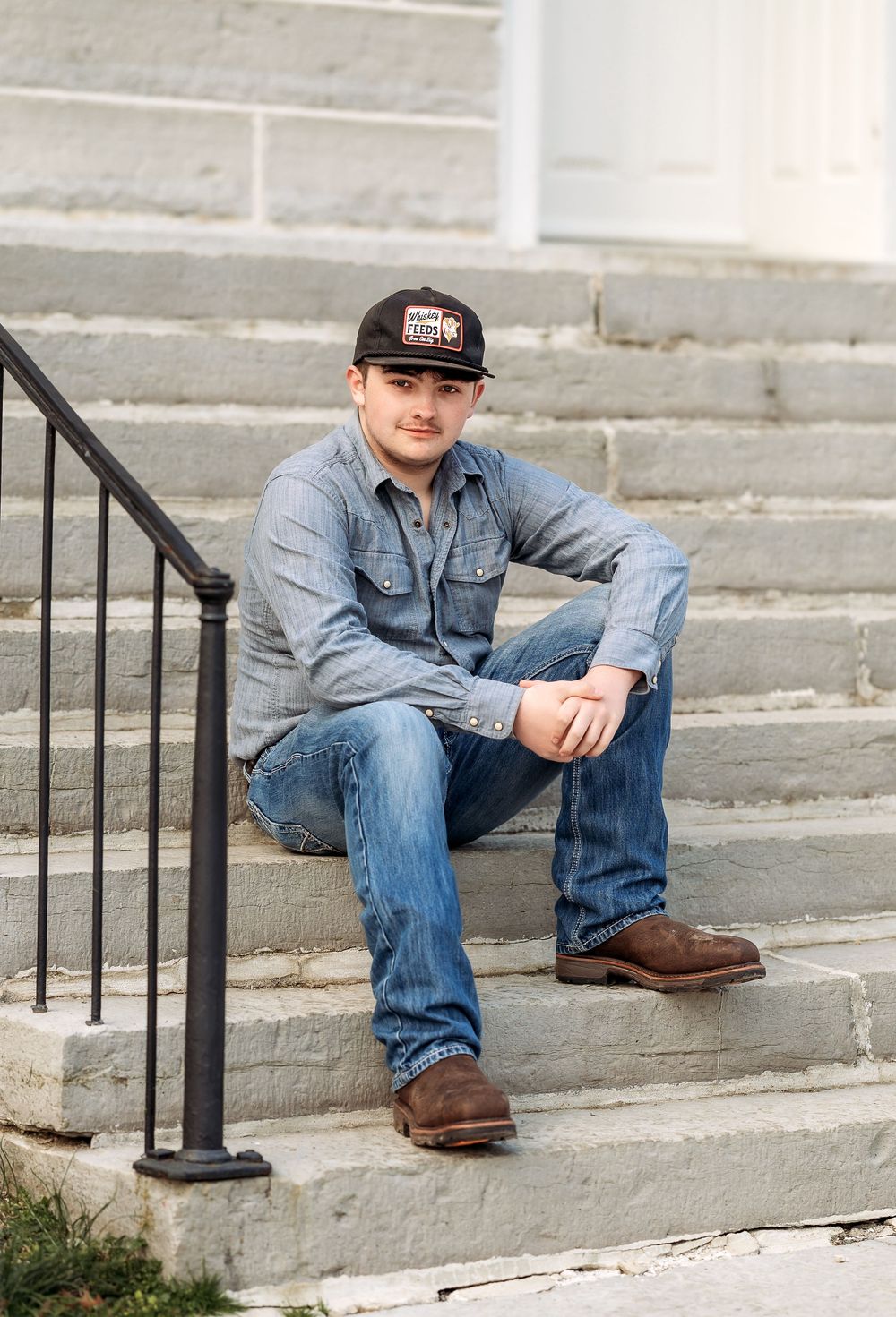 Someone sits casually on concrete steps wearing a denim jacket, jeans, baseball cap and brown boots.