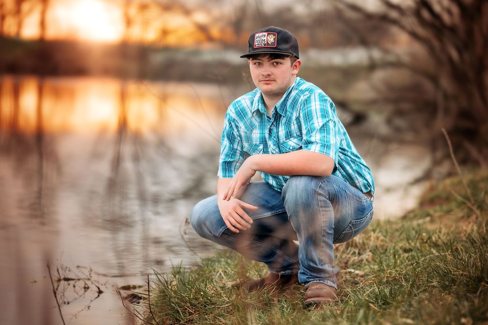 A person in blue plaid shirt and baseball cap crouches by a lake at sunset, creating a peaceful outdoor portrait.