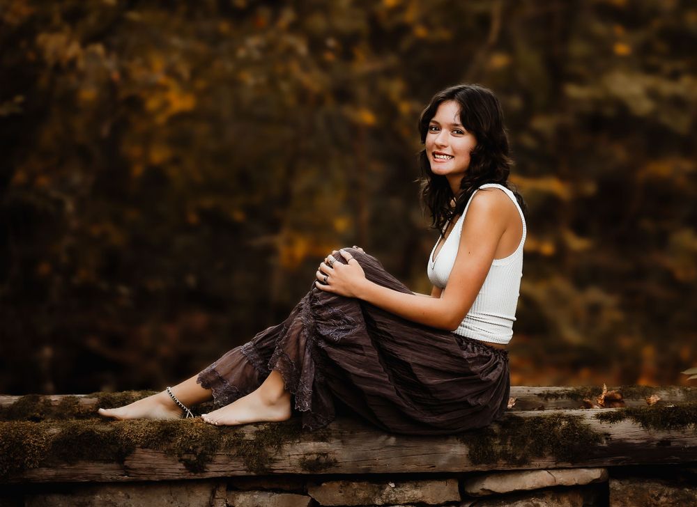 Woman in white tank top and flowy pants sits thoughtfully on a fallen log in autumn setting.