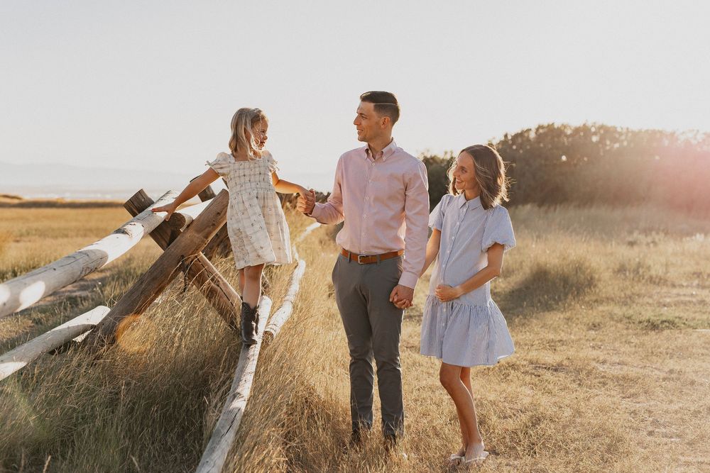 A father holds hands with two young daughters in a sunny golden field near a wooden fence.