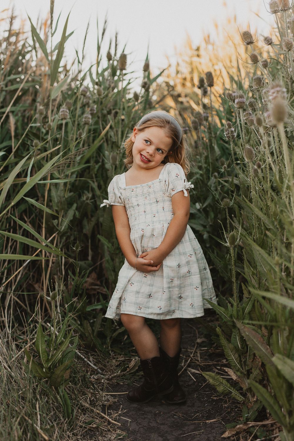 A young girl in a white floral dress and brown boots smiles while standing on a path surrounded by tall green plants.