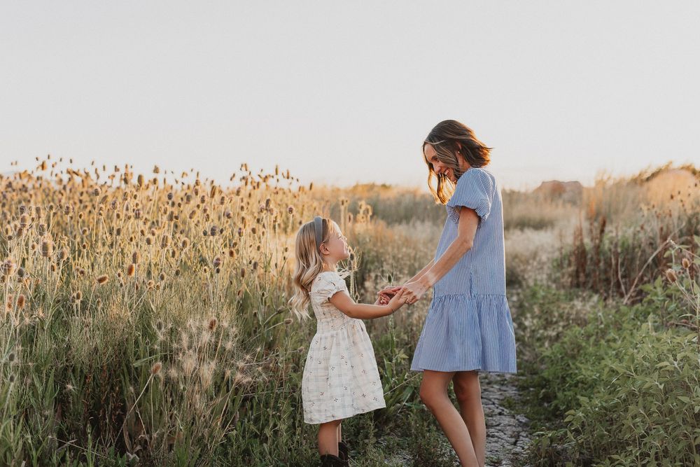 A mother and young daughter hold hands in a golden field of wildflowers during a warm sunset.