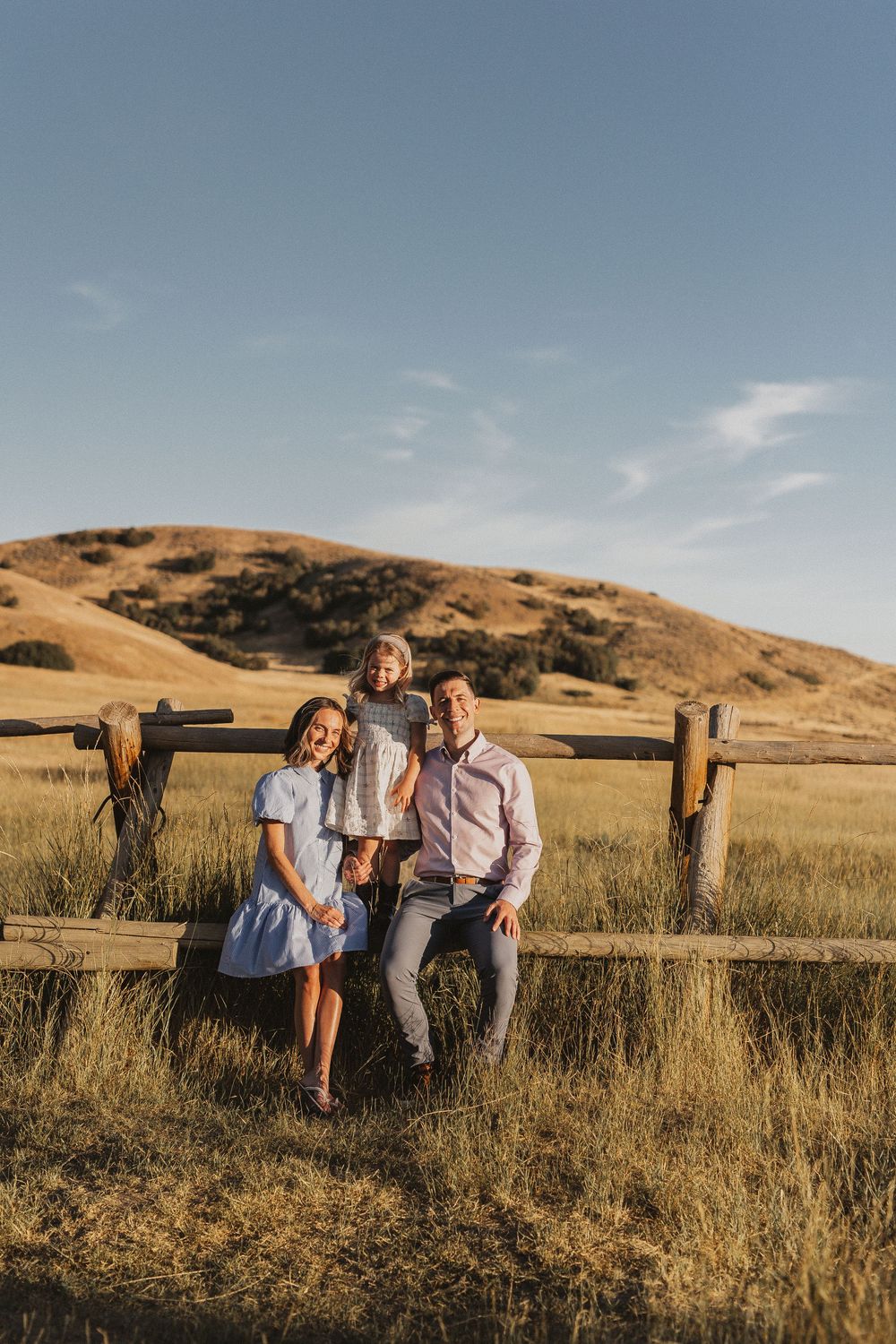 Family of three posing by a rustic wooden fence in a golden field with rolling hills under a clear blue sky.