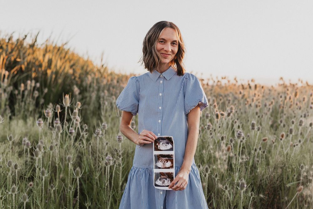 Smiling pregnant woman in blue dress holding ultrasound photos in a lavender field at golden hour.