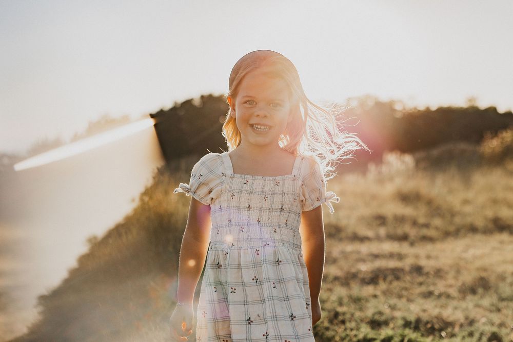 A young girl in a floral dress smiles joyfully outdoors in golden sunlight with her hair blowing in the wind.