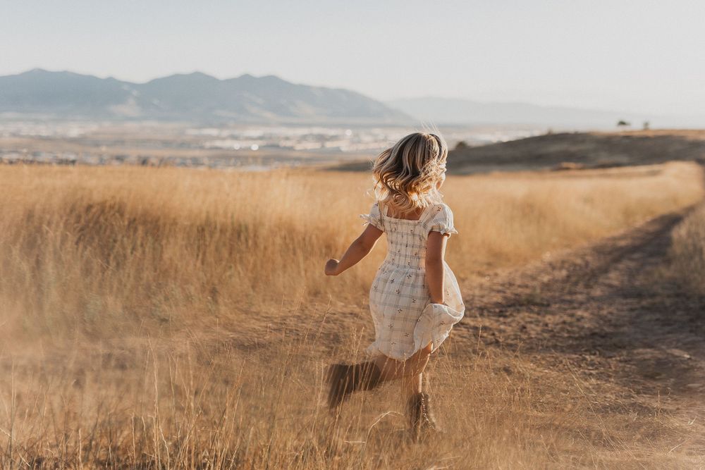 A young girl in a white dress runs through golden fields with mountains in the background on a sunny day.